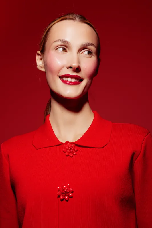 Young woman in red sweater smiles looking sideways against red background. Studio portrait with bright makeup and smooth skin. Concept of fashion, beauty, and confident female model.