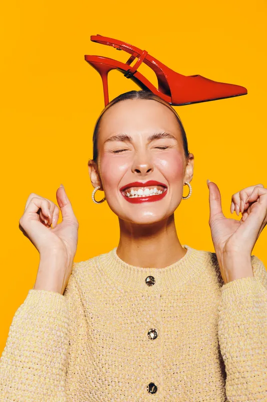 Happy woman with red lipstick and hoop earrings smiles with closed eyes, balancing red high heel shoe on head against bright yellow background. Concept of fun and fashion.