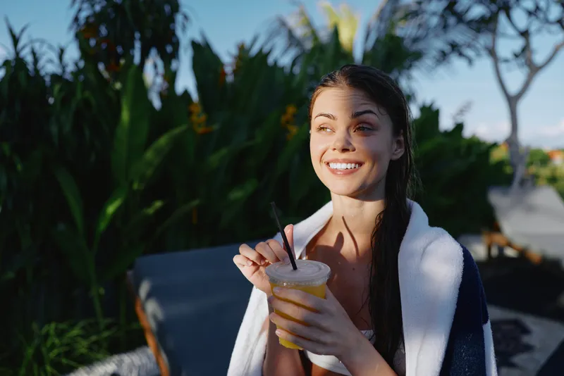 Happy woman outdoor with towel over shoulders drinks a smoothie, enjoying tropical scenery and summer leisure, bright smile amid lush plants, casual lifestyle under sunny daylight, healthy