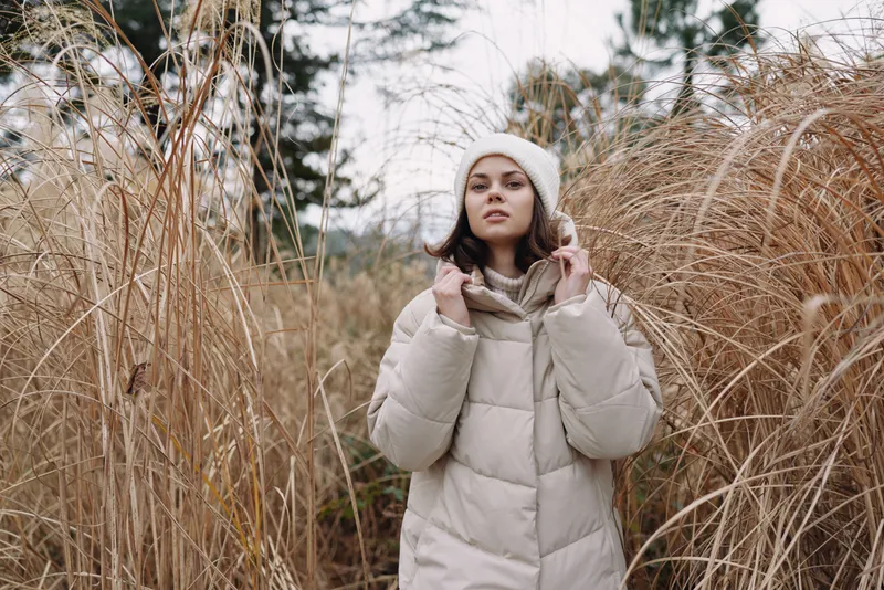 Woman in a beige winter coat and knit hat stands among tall grasses in a cold outdoor setting, exuding calm and style today
