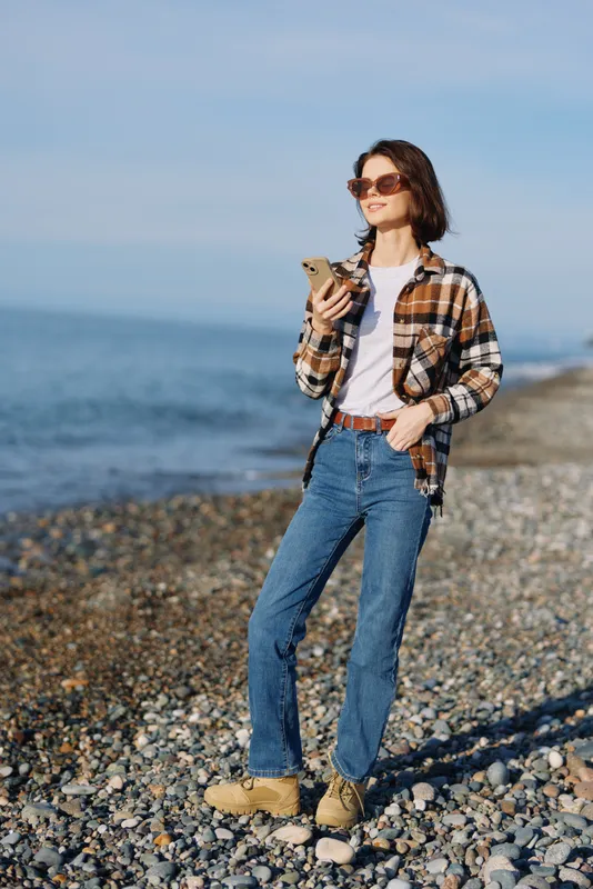 Woman in casual outdoor attire strolling on a rocky beach, holding a smartphone, wearing sunglasses, flannel shirt and jeans, enjoying a sunny day by the sea.