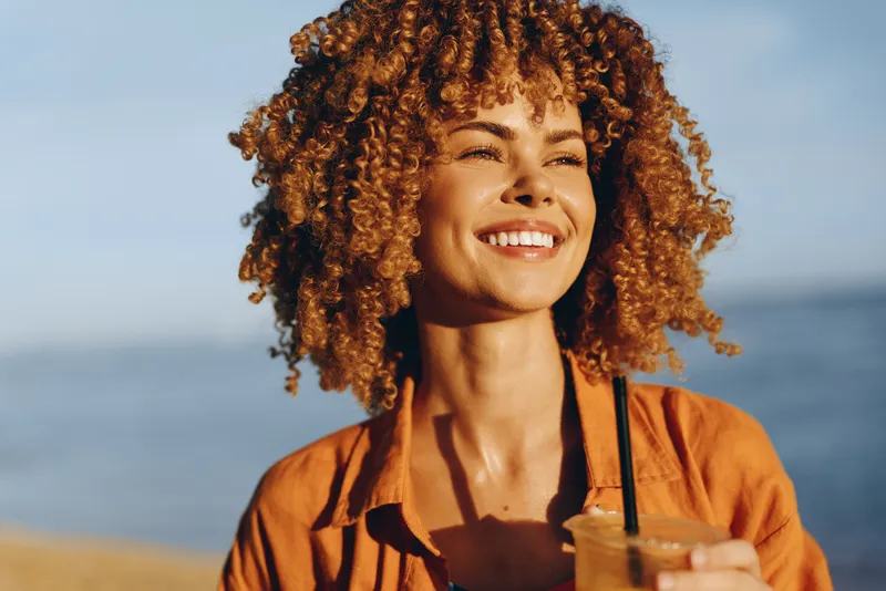 Woman curly hair smiling beach ocean vacation orange shirt drink summer portrait, young woman at the shoreline, warm sunlight and blue sky, holding a drink cup, relaxed travel mood