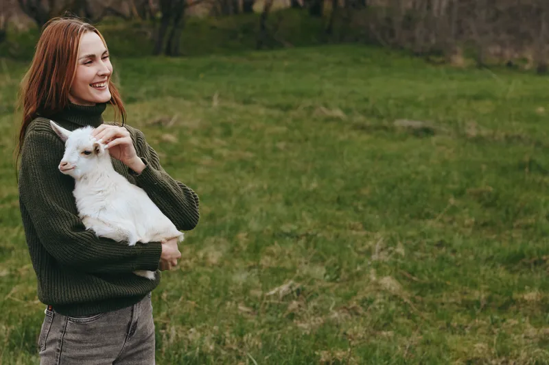 A woman in a dark sweater holds a white goat kid in a grassy field, smiling softly. A warm lifestyle moment emphasizing bond between person and farm animal outdoors.