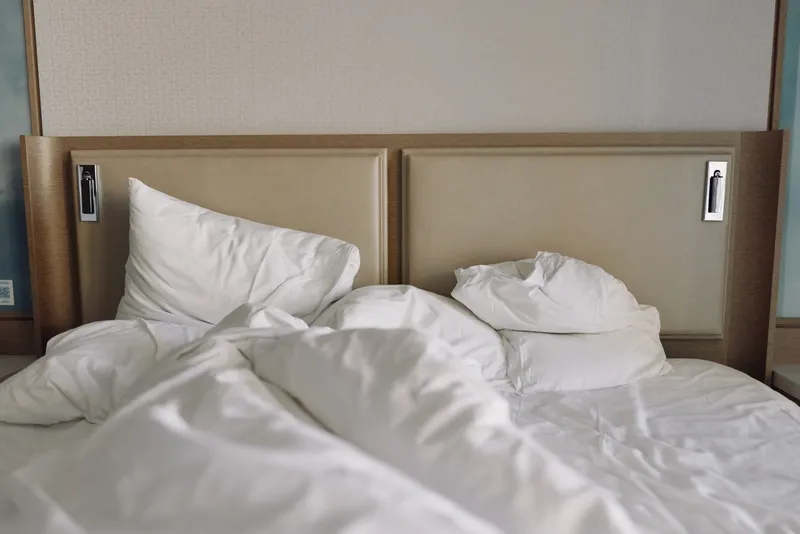 Unmade bed with white sheets and fluffy pillows, rumpled duvet creating a cozy, calm bedroom scene with soft light and inviting comfort