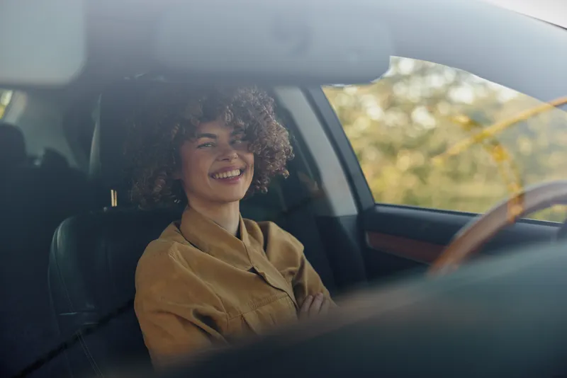 A smiling woman sits in a car wearing a brown jacket, captured in a candid lifestyle moment. Warm retro film color tones enhance the relaxed everyday scene, bright sunlit.