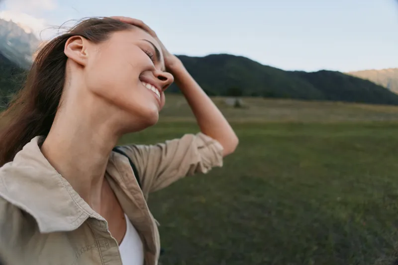 A happy woman in casual clothes enjoys nature outdoors with eyes closed and hand on head, surrounded by green fields and mountains in the background.