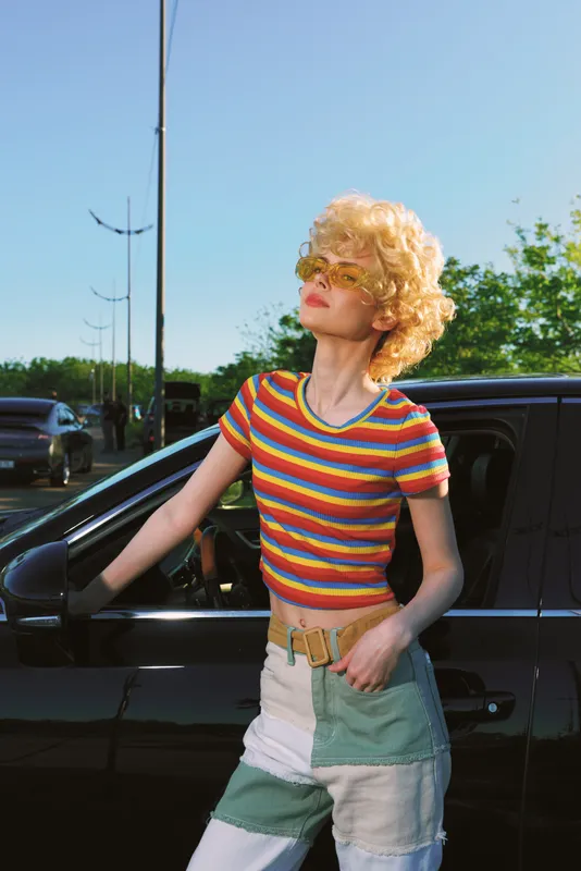 Woman wearing a retro striped top and patchwork shorts poses beside a car, showcasing casual lifestyle and vintage fashion, sunny outdoor scene with bright colors and wind in her hair