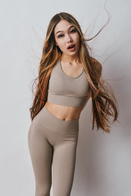 Young woman with long hair wearing beige sportswear posing against white background. Fitness girl in seamless activewear, casual posture with loose hair and natural expression in studio.