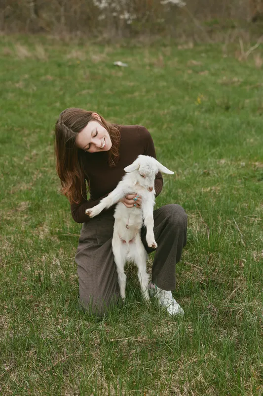 Young woman in a cozy sweater kneels in a grassy field, holding a small goat kid with tenderness, capturing a warm lifestyle moment of bonding between human and animal outdoors