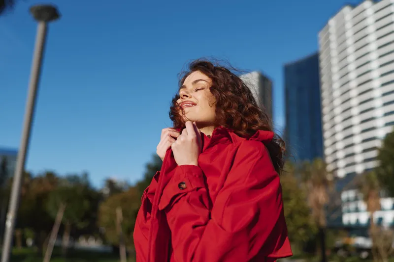 A woman in a red coat basks in bright sun, eyes closed and head tilted back. An urban backdrop of tall buildings frames a relaxed, confident portrait.