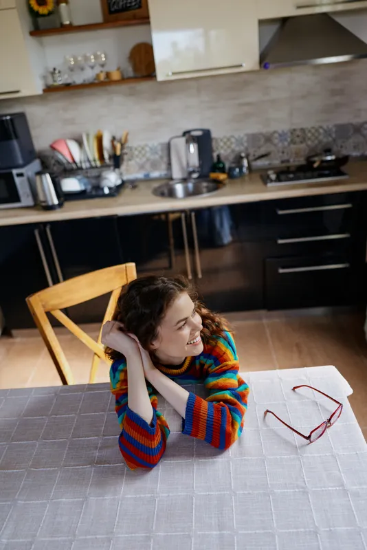Young woman with curly hair rests her head on her hands at a bright kitchen table, wearing a colorful striped sweater and glasses nearby.
