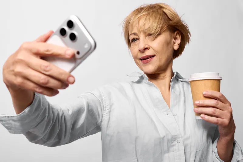 Woman senior taking a selfie with smartphone, holding coffee cup. White studio portrait, soft light, friendly smile, casual morning routine, communication and connection.
