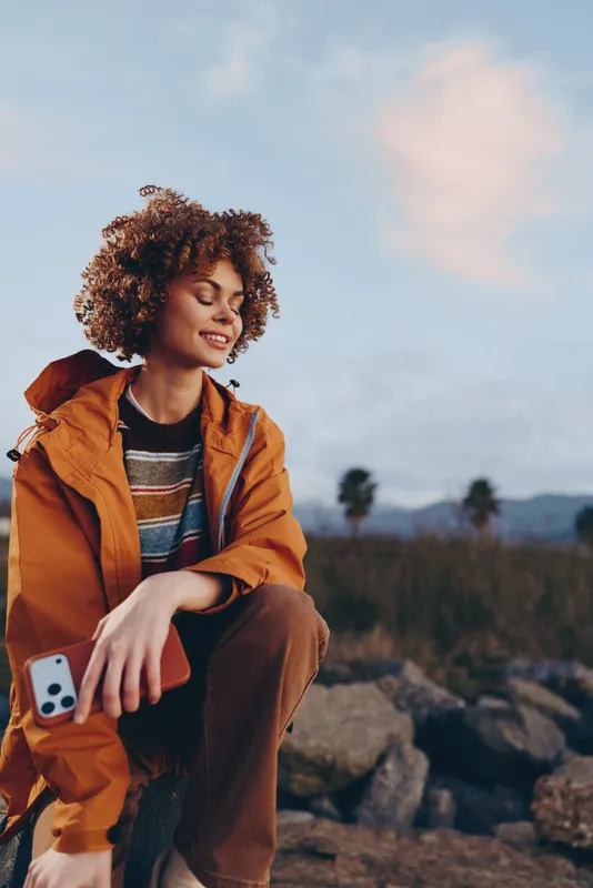 Woman lifestyle outdoors portrait, curly hair, orange jacket and phone in hand, sitting on rocky landscape, relaxed smile, warm sunlight, nature and wide sky background.