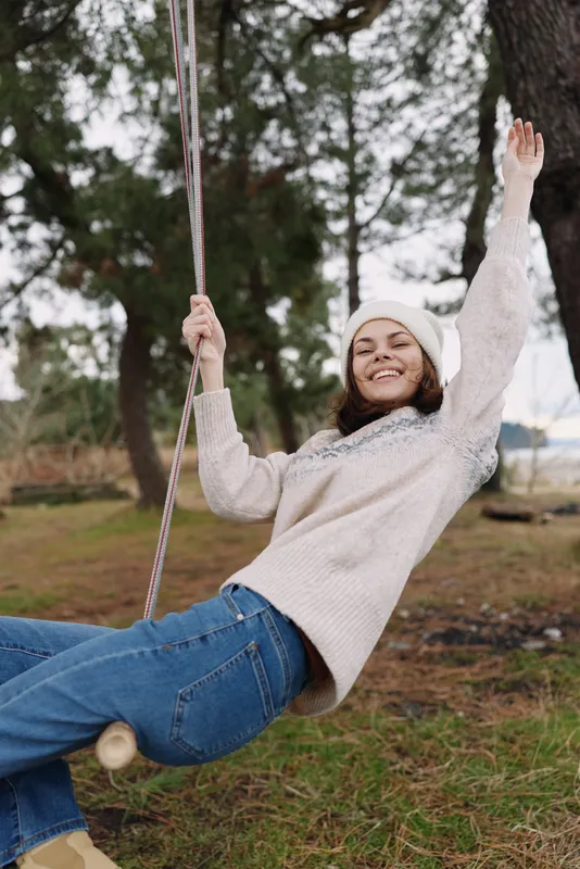 Girl on a swing smiling outdoors in a cozy sweater and beanie, enjoying a playful moment in a park with trees and open space on a sunny day.