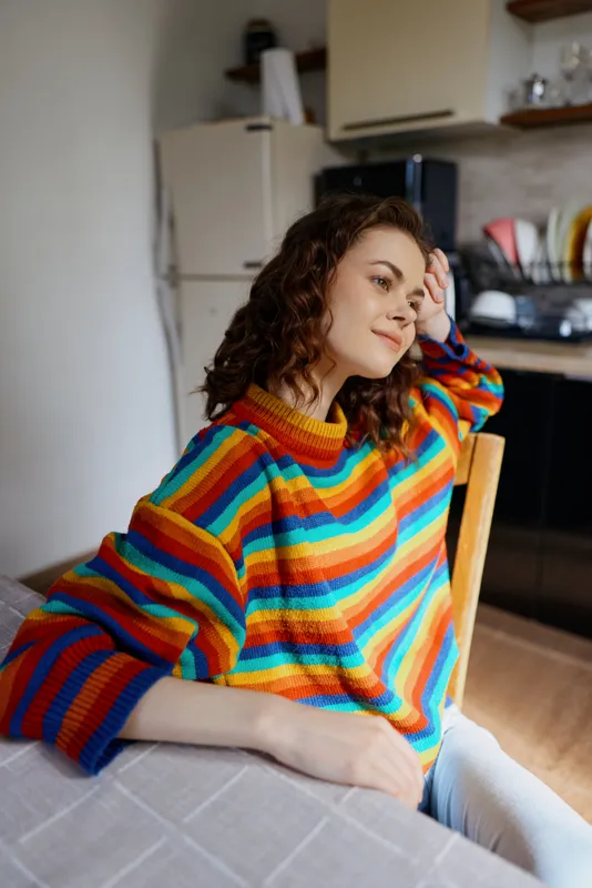 A woman in a colorful striped sweater sits at a home table, looking contemplative with a hand on her head. A cozy kitchen space frames the scene, conveying reflective mood and everyday comfort.