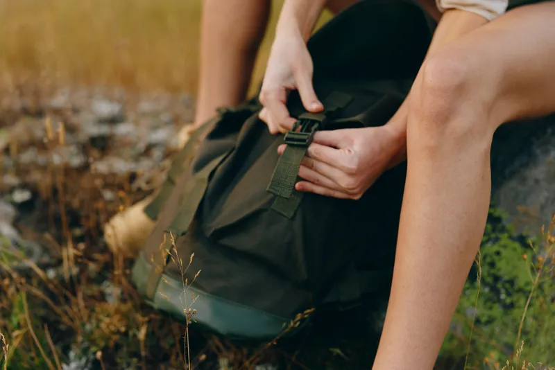 A young woman sits on grass outdoors, fastening a backpack strap with calm focus, ready for a relaxed hike in a natural setting.