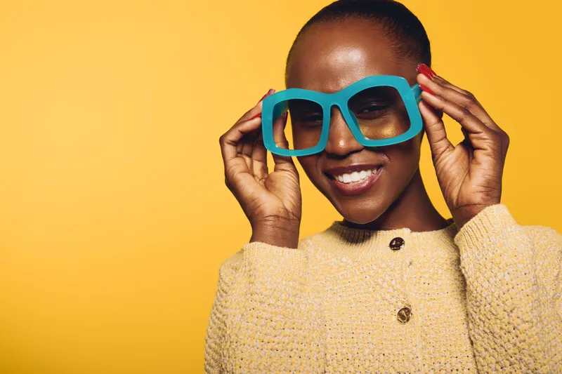 Smiling dark skinned African woman with short hair wearing oversized blue glasses and beige sweater poses against yellow background. Concept of fashion, joy, style, and confident beauty.