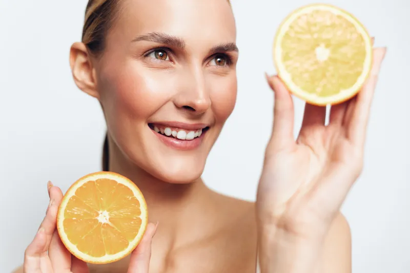 Smiling woman closeup holding orange slices for skincare beauty portrait, bright studio light, fresh citrus concept, smooth skin, relaxed gaze, soft background fresh glow