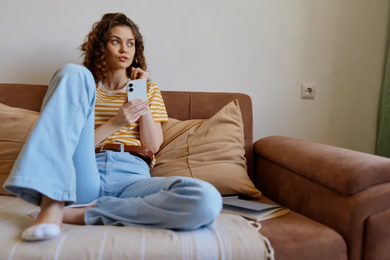 A young woman in a striped yellow tee and light jeans sits on a beige sofa at home, scrolling on her phone with a relaxed, focused expression amid soft cushions and natural light.