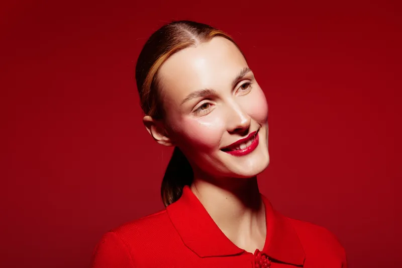 Smiling woman with red lipstick and red sweater poses against red background. Studio portrait with bright makeup and warm lighting. Concept of beauty and fashion.