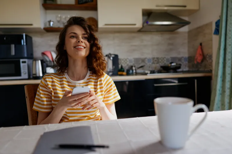 Young woman with curly hair in a striped t shirt sits at a kitchen table, glancing at her smartphone with a cheerful smile, a warm, casual home moment illuminated by soft morning light and modern
