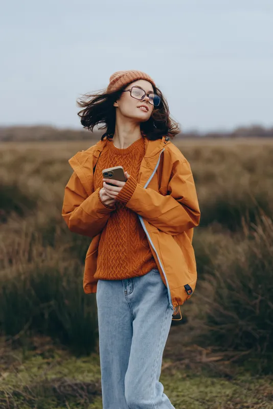 Woman lifestyle outdoors field, orange jacket and glasses, standing and holding a smartphone, looking aside, calm casual mood in nature, rural grass young portrait