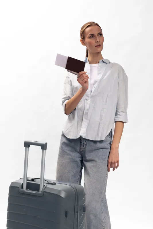 Young woman in casual clothes holding passport and boarding pass stands next to suitcase on white background. Concept of travel, vacation planning, and airport security check.