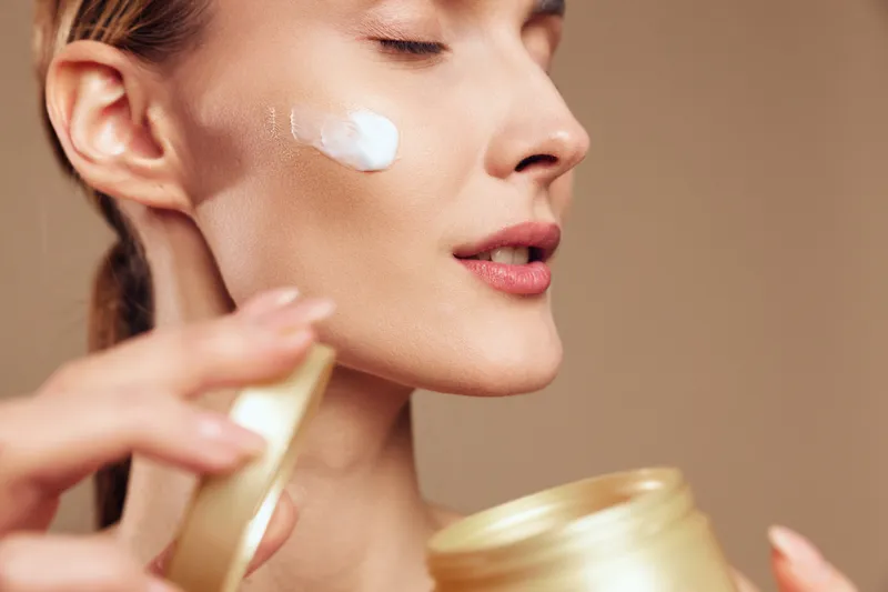Close up of a woman applying cream to her cheek, highlighting a skincare routine, smooth skin, and a soft warm glow on a neutral background, in a studio setting.
