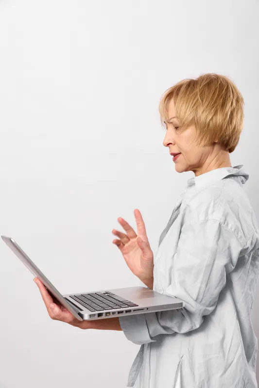 Woman with short blond hair holds laptop, waving hand in greeting, side view, online communication for call, focused work mood, white background isolation, studio portrait