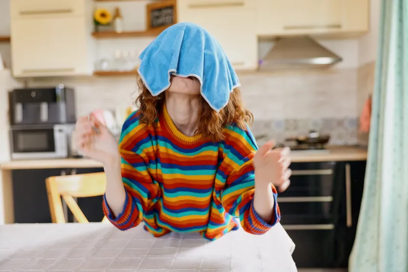 Playful woman at home covers her face with a towel after shower, wearing a bright striped shirt, in a sunny kitchen, capturing a casual, cozy lifestyle moment, candid and relatable