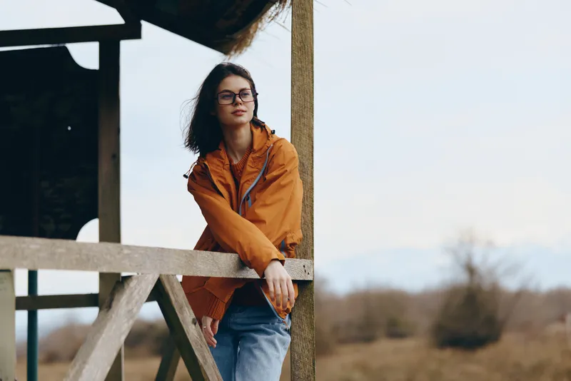 Woman lifestyle outdoors, wearing an orange jacket and glasses, leans on a wooden porch railing with a calm smile. Soft daylight and rural trees blur behind her, serene portrait.