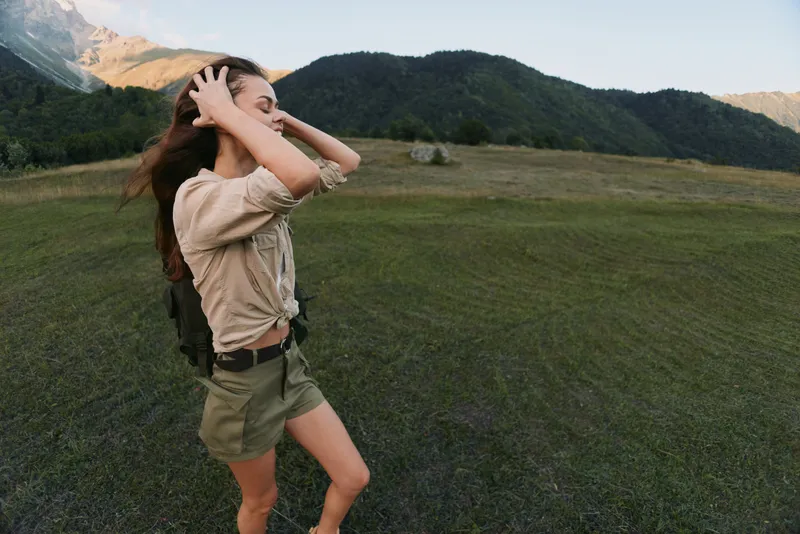 Woman hiking in a grassy valley, adjusting hair, wearing beige shirt and shorts, backpack ready, surrounded by mountains and wide open sky, vibrant outdoor lifestyle moment