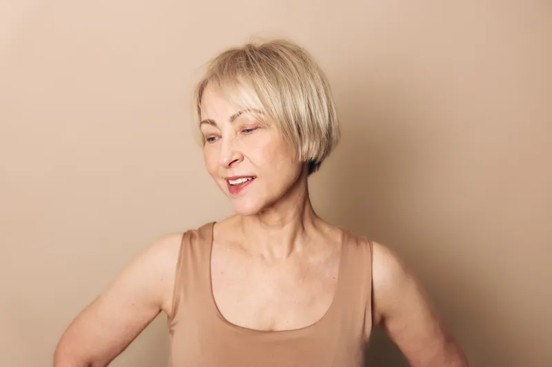 Smiling mature woman with short blonde hair wearing sleeveless beige top looks away thoughtfully on beige background. Concept of aging beauty, confidence, and natural skin care for older adults.