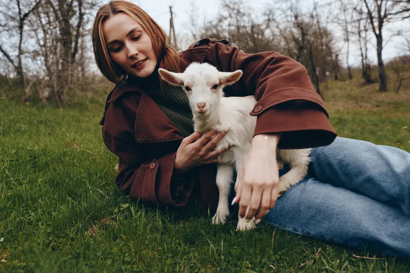 A young woman lies on a grassy field, cradling a white goat kid in her arms, capturing a tender moment of outdoor lifestyle, animal companionship, and calm countryside atmosphere.
