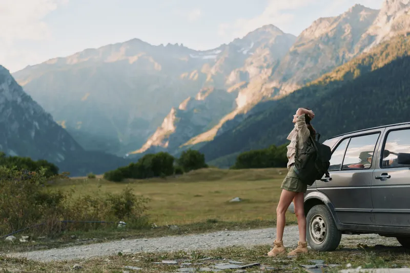 Woman traveler by a rugged mountain road stands beside a car, wearing casual jacket and shorts, enjoying open air, a nature escape on a serene alpine valley road