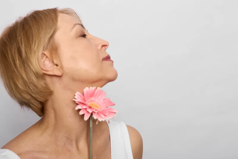 Woman profile in studio, pink flower near her neck, soft light and closeup beauty mood, gentle smile, smooth skin, minimal white background, calm spa feeling