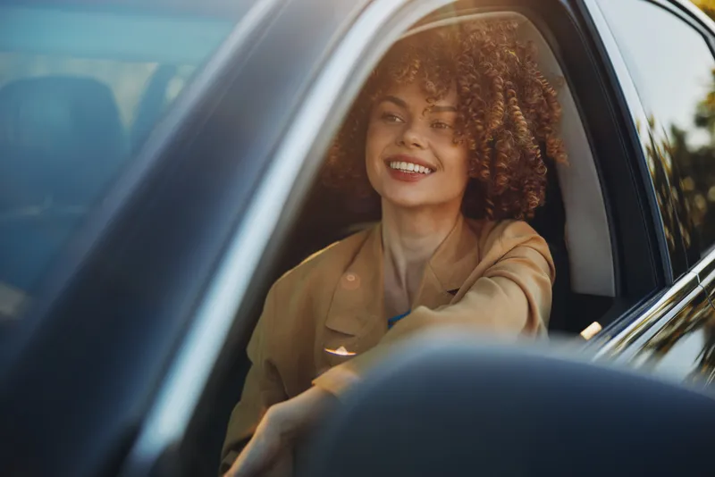 Joyful woman with curly hair sits in a car, wearing a light brown jacket, enjoying a casual lifestyle moment with retro film color tones, smiling toward the camera.