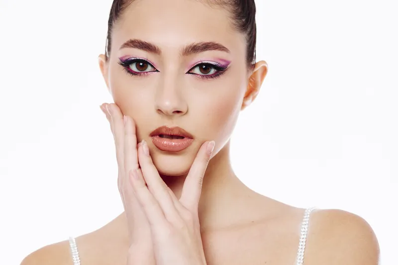 Young woman with bold pink and black eye makeup poses sensually on a white background. Hands gently touch her face highlighting flawless skin in fashion beauty concept.