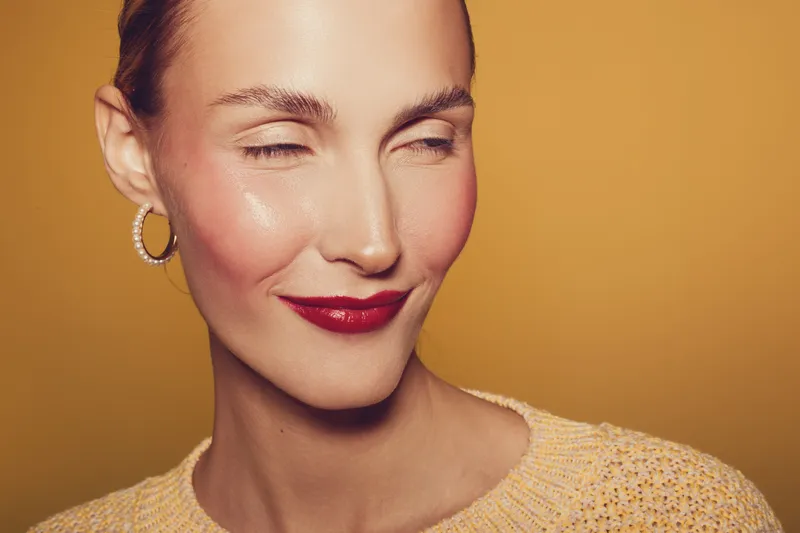Close-up of a smiling woman with red lipstick and hoop earrings wearing a yellow sweater on a warm yellow background. Concept of beauty, fashion, and confident expression.