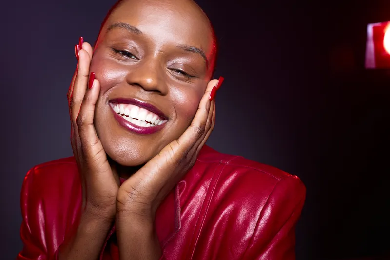 Close-up of a happy African woman with dark skin wearing a red leather jacket smiling with hands on face. Studio lighting highlights her joyful expression and smooth skin tone.