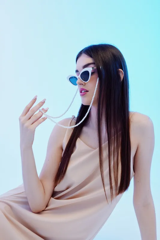 Young woman with long dark hair wearing stylish sunglasses and a beige dress holds a pearl chain. Studio shot on a light blue background. Concept for woman beauty fashion.