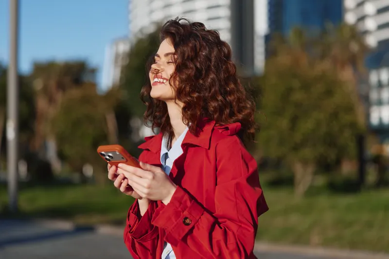 Woman in a red coat smiles while holding a smartphone outdoors, sunny park setting, urban skyline in the background, casual style and cheerful mood.