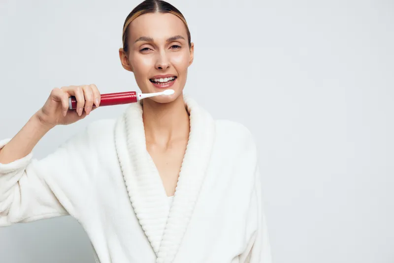 Smiling woman in white bathrobe brushes teeth with electric toothbrush, showing dental care routine, clean teeth concept, isolated on light background.