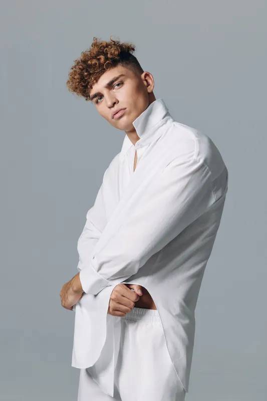 Young man with curly hair wearing white shirt and pants posing with arms crossed against gray background in studio for fashion portrait and style concept.