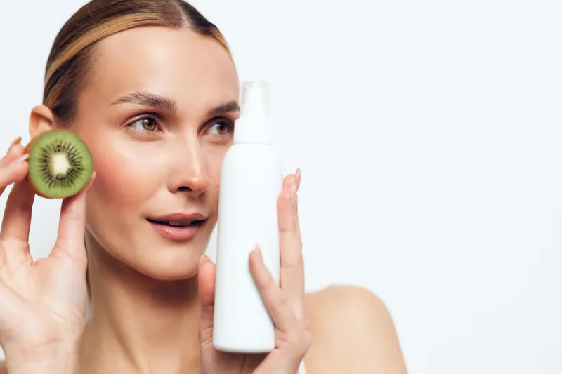 A young woman with clear skin holds a white cosmetic bottle and a sliced kiwi near her face. The clean background highlights skincare and natural beauty concepts.