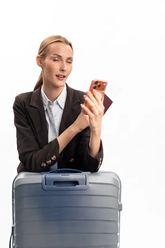 Young businesswoman with suitcase checks phone while holding passport, dressed in formal jacket and shirt. Concept of travel planning, business trip, and modern lifestyle.