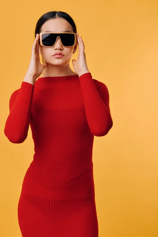 Woman in red fitted dress and large black sunglasses posing on yellow background. Fashion model with long black hair in studio, holding glasses frame for style and attitude portrait.
