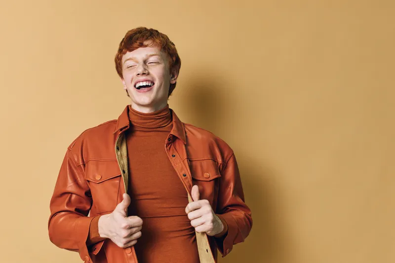 Young man with red hair and freckles wearing orange jacket and turtleneck smiling with eyes closed showing thumbs up gesture on beige background in studio portrait.