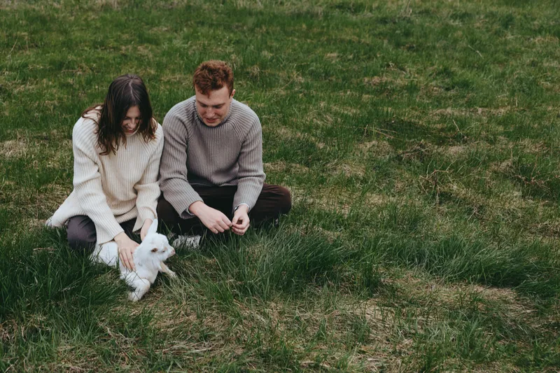 A couple sits on a grassy meadow with a goat kid, enjoying a calm outdoor moment, a wholesome lifestyle scene that highlights family, nature, companionship, and simple rural joy.