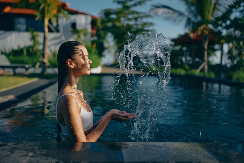 Woman in a white swimsuit stands in a pool, water arcing from a fountain and glistening in sunlight. A serene moment of relaxation, outdoor leisure, and tropical wellness at a resort.